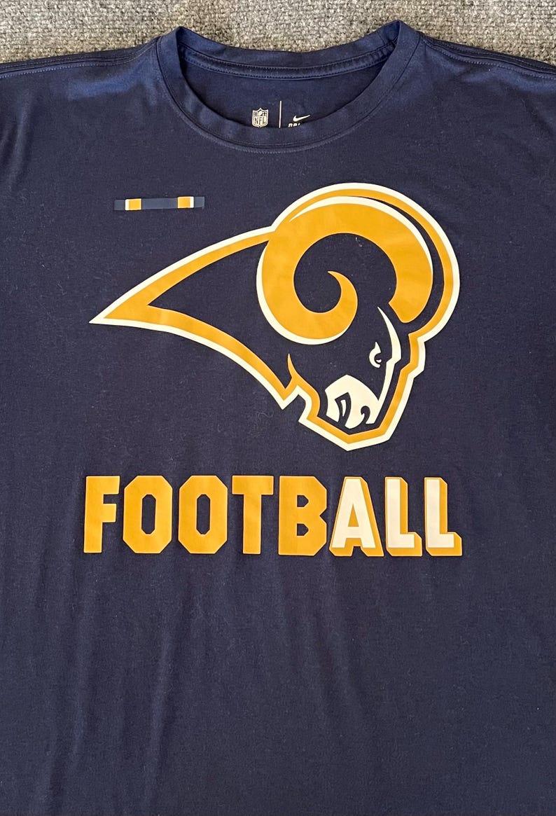 Op de afbeelding: Marineblauw T-shirt met het Los Angeles Rams logo in goud en wit, en het woord "FOOTBALL" in gouden blokletters. Het shirt heeft een ronde hals en korte mouwen. Het NFL-logo is zichtbaar.