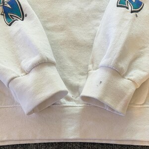 Peut inclure: Un sweat-shirt blanc &agrave; manches longues avec des poignets et un ourlet c&ocirc;tel&eacute;s. Les manches pr&eacute;sentent un motif graphique avec une lettre N en d&eacute;grad&eacute; bleu et sarcelle. Une petite tache sombre est visible sur le poignet droit pr&egrave;s du bord.