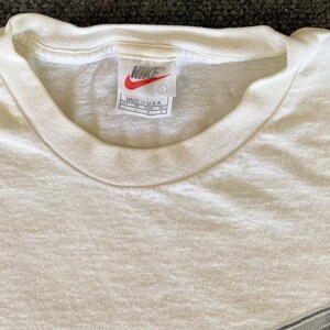 May include: Cream-colored, short-sleeved t-shirt with a crew neck. The Nike logo is visible on the tag, along with the text "MADE IN U.S.A." The shirt is a size large.