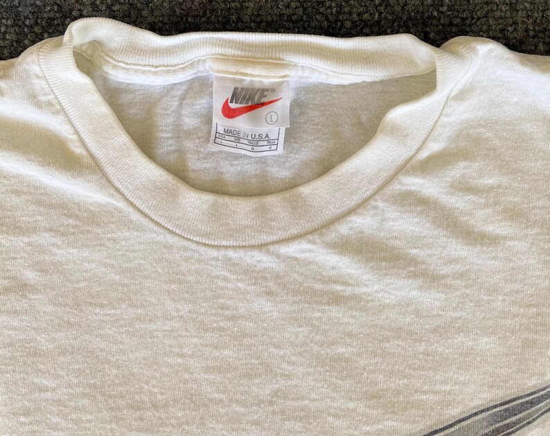 May include: Cream-colored, short-sleeved t-shirt with a crew neck. The Nike logo is visible on the tag, along with the text "MADE IN U.S.A." The shirt is a size large.