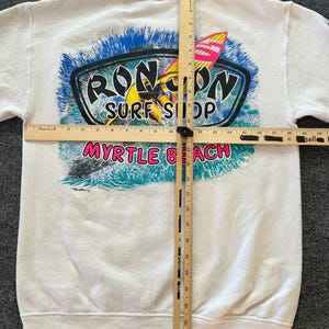 Peut inclure: Un sweat-shirt blanc &agrave; manches longues avec un motif de surfeur sur une vague et le texte RON JON SURF SHOP MYRTLE BEACH. Deux r&egrave;gles en bois sont pos&eacute;es sur le v&ecirc;tement, indiquant une largeur de 50,8 cm et une longueur de 66 cm.