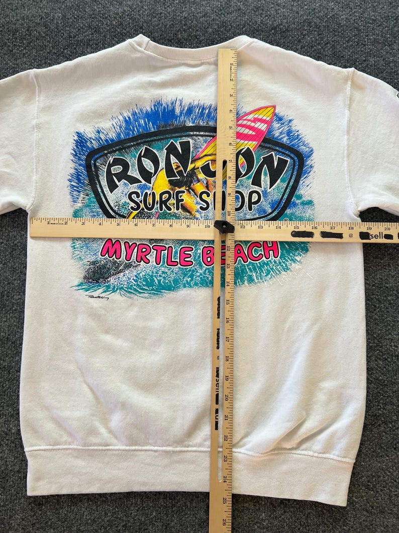 Peut inclure: Un sweat-shirt blanc &agrave; manches longues avec un motif de surfeur sur une vague et le texte RON JON SURF SHOP MYRTLE BEACH. Deux r&egrave;gles en bois sont pos&eacute;es sur le v&ecirc;tement, indiquant une largeur de 50,8 cm et une longueur de 66 cm.