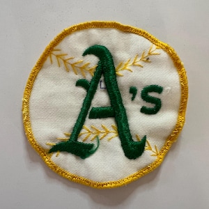 May include: A circular embroidered patch featuring a white background with yellow stitched baseball-style laces. The center displays the green embroidered text A's in a stylized font. The entire patch is bordered by a thick yellow stitched edge.