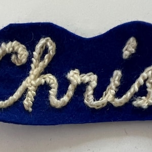 May include: A royal blue felt patch featuring the name Chris embroidered in white yarn. The lettering is executed in a cursive script style, with the white yarn standing out against the solid blue fabric background of the patch.
