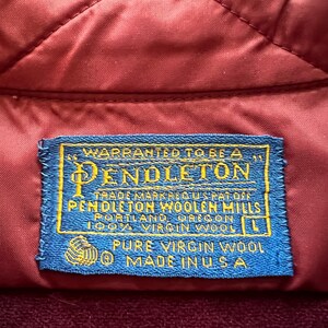 May include: A rectangular blue fabric clothing label with yellow text stitched onto a quilted maroon garment. The label reads: WARRANTED TO BE A PENDLETON, TRADE MARK REG US PAT OFF, PENDLETON WOOLEN MILLS, PORTLAND, OREGON, 100% VIRGIN WOOL, L, PURE VIRGIN WOOL, MADE IN USA.