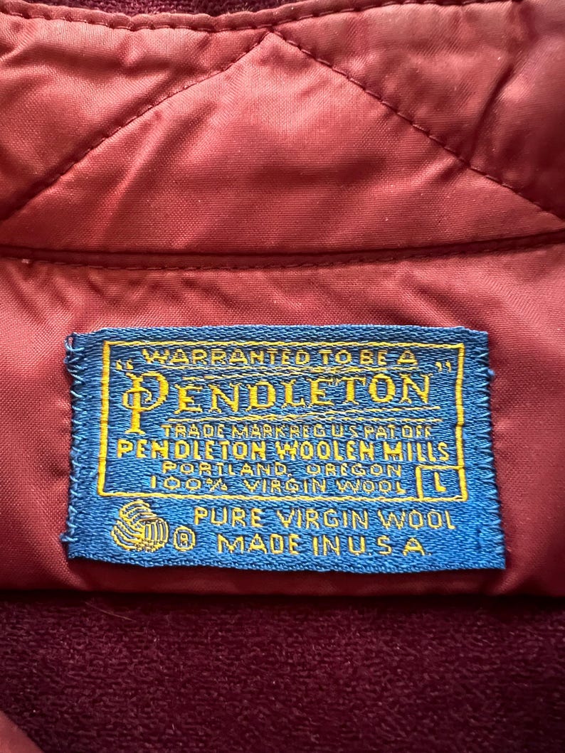 May include: A rectangular blue fabric clothing label with yellow text stitched onto a quilted maroon garment. The label reads: WARRANTED TO BE A PENDLETON, TRADE MARK REG US PAT OFF, PENDLETON WOOLEN MILLS, PORTLAND, OREGON, 100% VIRGIN WOOL, L, PURE VIRGIN WOOL, MADE IN USA.