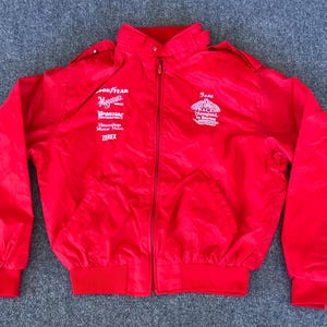 May include: A bright red racing jacket with a zip-up front, collar, and elastic cuffs and waistband. The jacket features white logos for Goodyear, Magness, Pontiac, and Zerex. The text "I am RACER Disneyland in Boston" is also visible.