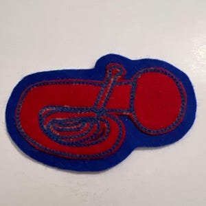 May include: A felt patch featuring a red tuba shape outlined in blue thread on a blue background. The design shows the brass instrument's coiled tubing and large bell, with the blue felt base cut to follow the contour of the red tuba shape.