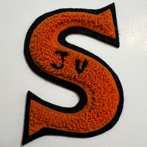 May include: A varsity-style letter S patch featuring a textured orange chenille center with a black felt border. The letter S has the black embroidered letters JV positioned in the center of the orange fabric, creating a layered appearance.