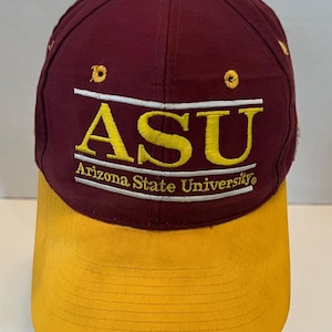 May include: A maroon baseball cap with a yellow brim and a yellow button on top. The front features embroidered yellow text that reads ASU and Arizona State University, framed by two horizontal white lines and two yellow eyelets above the text.