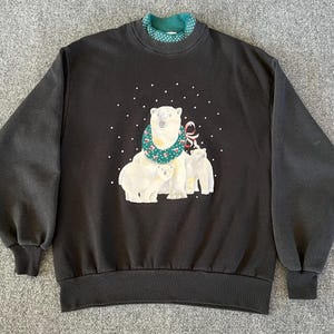 May include: Black sweatshirt featuring a polar bear family graphic. The polar bears are white with a green and white floral scarf. The sweatshirt has a green and white polka dot collar and white snowflake accents.