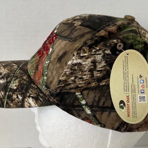 May include: A camouflage baseball cap with a curved brim. The cap features a brown, green, and tan pattern, with the word "Mossy Oak" on a hang tag. The cap is designed for outdoor activities.