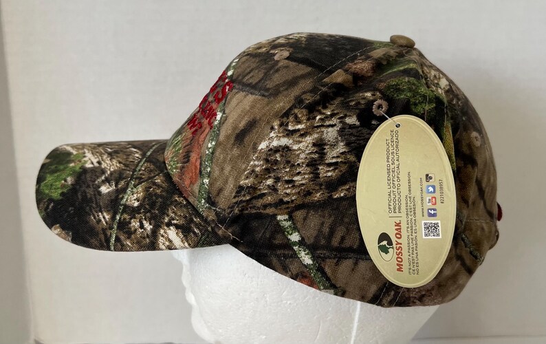 May include: A camouflage baseball cap with a curved brim. The cap features a brown, green, and tan pattern, with the word "Mossy Oak" on a hang tag. The cap is designed for outdoor activities.