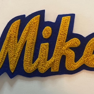 May include: A custom name patch featuring the name Mike in yellow chenille embroidery with a textured, looped surface. The letters are outlined and set against a solid dark blue felt backing that follows the contour of the script lettering.