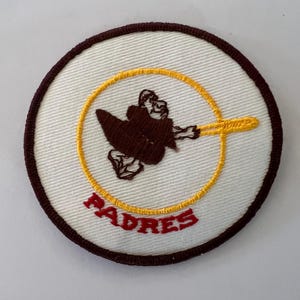 May include: A circular embroidered patch featuring a white background with a dark brown border. In the center, a brown monk figure is depicted inside a yellow circular outline, with the red text PADRES embroidered below the figure.
