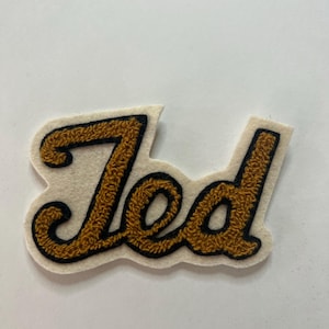 May include: A sew-on patch featuring the name Ted in gold-colored chenille embroidery with a black outline. The letters are set against a cream-colored felt backing that follows the contour of the script lettering in a cursive font style.