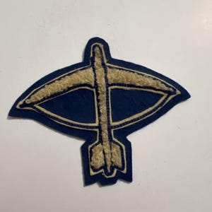 May include: A navy blue fabric patch featuring a textured, cream-colored embroidered design of a crossbow. The patch has a distinct, pointed shape that follows the outline of the weapon, with the cream embroidery highlighting the bow and arrow.