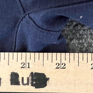 Op de afbeelding: Close-up van marineblauwe stof met zichtbare stiksels. Een houten liniaal met zwarte markeringen is over de stof geplaatst en toont metingen. De markeringen van de liniaal zijn in centimeters.