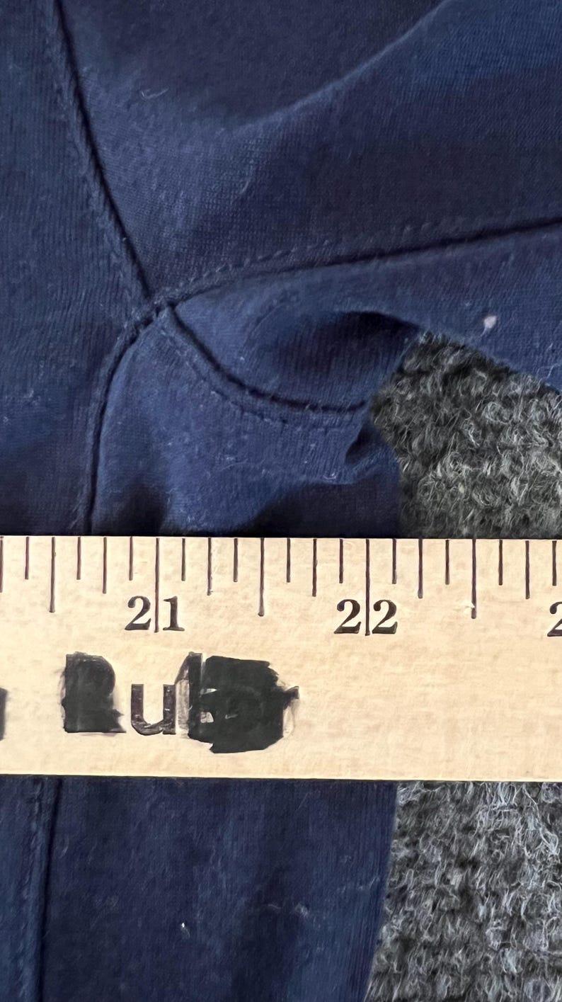 Op de afbeelding: Close-up van marineblauwe stof met zichtbare stiksels. Een houten liniaal met zwarte markeringen is over de stof geplaatst en toont metingen. De markeringen van de liniaal zijn in centimeters.