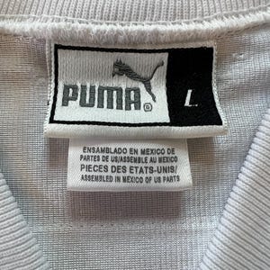 Op de afbeelding: Close-up van het label van een wit sportshirt. Het label toont het PUMA-logo en het woord "PUMA" in zwart. Daaronder staat "ASSEMBLED IN MEXICO OF US PARTS". De maat is "L".