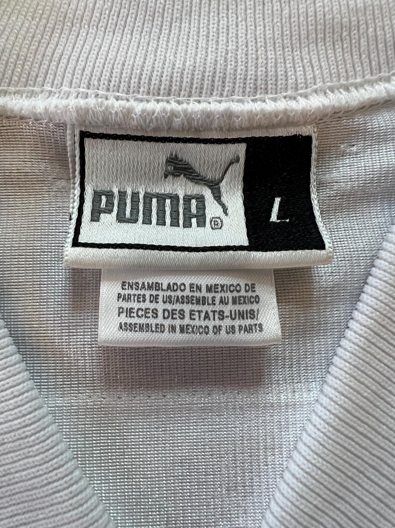 Op de afbeelding: Close-up van het label van een wit sportshirt. Het label toont het PUMA-logo en het woord "PUMA" in zwart. Daaronder staat "ASSEMBLED IN MEXICO OF US PARTS". De maat is "L".