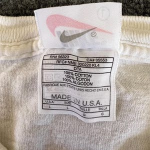 May include: Close-up of a white t-shirt tag with the Nike logo. The tag includes text such as "100% Cotton" and "Made in U.S.A." with size information. The tag is rectangular with rounded corners.