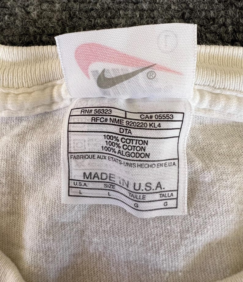 May include: Close-up of a white t-shirt tag with the Nike logo. The tag includes text such as "100% Cotton" and "Made in U.S.A." with size information. The tag is rectangular with rounded corners.