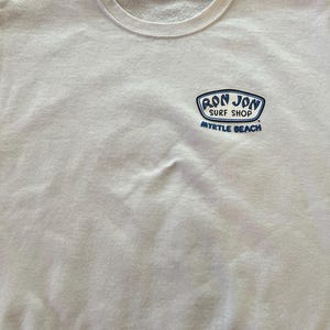 Peut inclure: Un sweat-shirt blanc &agrave; col rond avec un petit motif sur la poitrine gauche. Le motif est un embl&egrave;me bleu et blanc avec le texte RON JON SURF SHOP et MYRTLE BEACH imprim&eacute; en lettres bleues sous l'embl&egrave;me sur le tissu blanc du v&ecirc;tement.