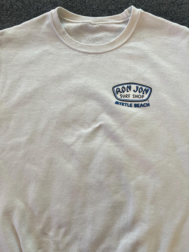 Peut inclure: Un sweat-shirt blanc &agrave; col rond avec un petit motif sur la poitrine gauche. Le motif est un embl&egrave;me bleu et blanc avec le texte RON JON SURF SHOP et MYRTLE BEACH imprim&eacute; en lettres bleues sous l'embl&egrave;me sur le tissu blanc du v&ecirc;tement.