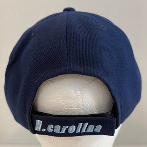 Pu&ograve; includere: Un cappello da baseball blu navy con visiera nera e un logo ricamato in blu chiaro che dice "N. Carolina".