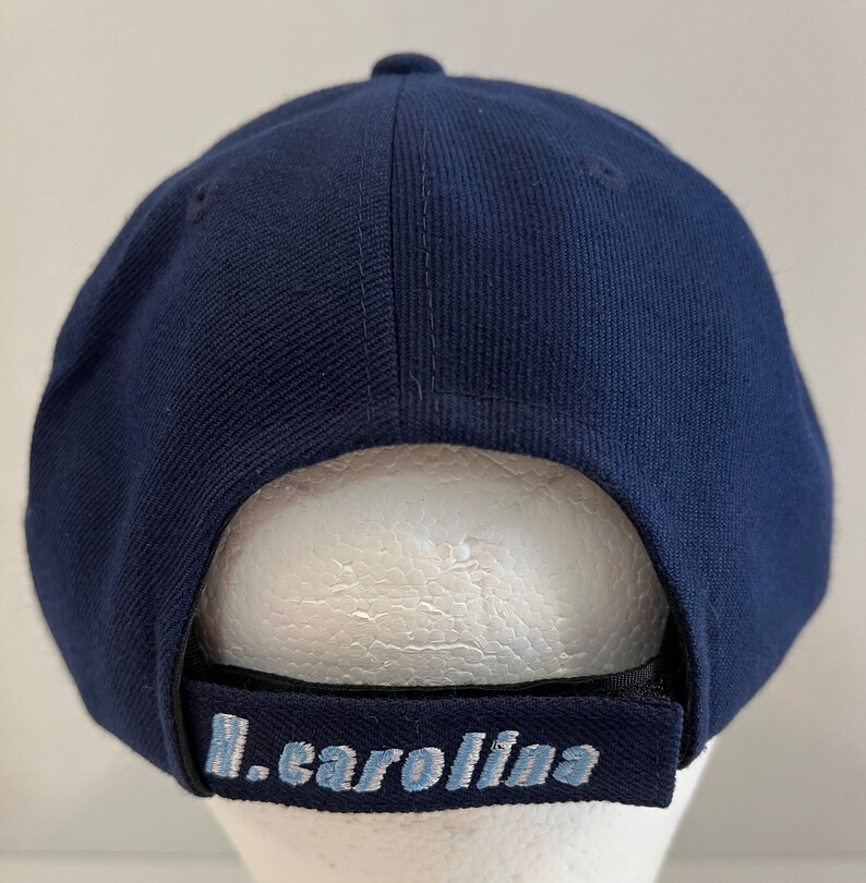 Pu&ograve; includere: Un cappello da baseball blu navy con visiera nera e un logo ricamato in blu chiaro che dice "N. Carolina".