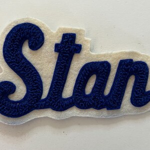 May include: A white felt patch featuring the name Stan embroidered in a dark blue, textured, cursive script. The lettering has a raised, looped stitch pattern that stands out against the smooth, off-white felt backing, which follows the contour of the text.