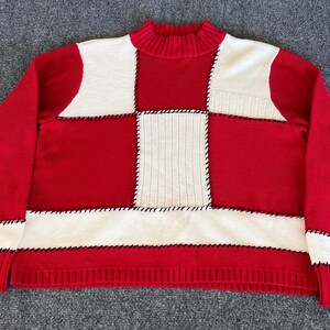 May include: A red and white patchwork sweater with a mock neck. The sweater features a checkerboard design with black stitching. The sleeves and bottom hem are ribbed. The sweater is made of a soft knit material.