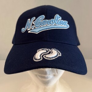 Pu&ograve; includere: Un cappello da baseball blu navy con un design ricamato bianco e azzurro che dice "N. Carolina". Il cappello ha anche un design ricamato bianco di due impronte di piedi sul davanti.