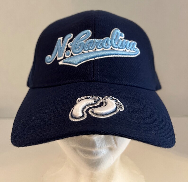 Pu&ograve; includere: Un cappello da baseball blu navy con un design ricamato bianco e azzurro che dice "N. Carolina". Il cappello ha anche un design ricamato bianco di due impronte di piedi sul davanti.
