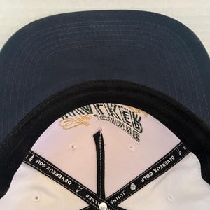 May include: A navy blue baseball cap with a white interior. The cap features the text "DEVEREUX GOLF" on the interior band and a logo with the text "WALKER" and "JOHN" on the interior. The cap has a black brim.
