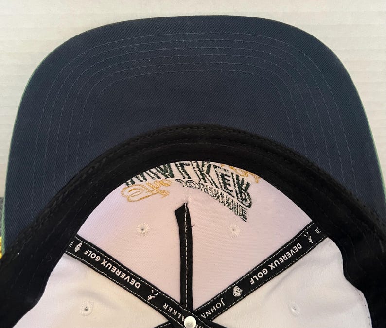 May include: A navy blue baseball cap with a white interior. The cap features the text "DEVEREUX GOLF" on the interior band and a logo with the text "WALKER" and "JOHN" on the interior. The cap has a black brim.