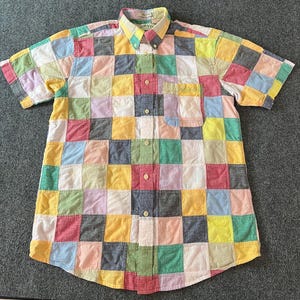 May include: A short-sleeve button-down shirt featuring a multi-colored patchwork pattern of squares in shades of yellow, pink, green, blue, and white. The shirt includes a single chest pocket, a button-down collar, and a front button placket.