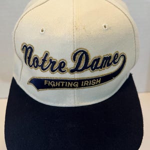 May include: A baseball cap with a cream-colored crown and a solid black brim. The front features embroidered text reading Notre Dame in navy blue with gold trim, and the words Fighting Irish in gold lettering on a navy blue banner.