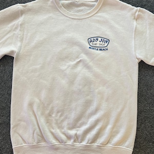Peut inclure: Un sweat-shirt blanc &agrave; manches longues et col rond, arborant un petit motif bleu et blanc sur la poitrine gauche. Le motif affiche le texte "RON JON SURF SHOP" et "MYRTLE BEACH" dans une police stylis&eacute;e. Le v&ecirc;tement poss&egrave;de un col c&ocirc;tel&eacute;.
