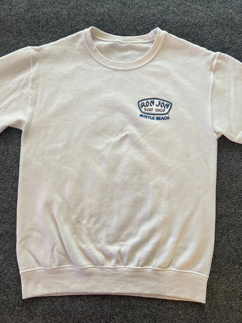 Peut inclure: Un sweat-shirt blanc &agrave; manches longues et col rond, arborant un petit motif bleu et blanc sur la poitrine gauche. Le motif affiche le texte "RON JON SURF SHOP" et "MYRTLE BEACH" dans une police stylis&eacute;e. Le v&ecirc;tement poss&egrave;de un col c&ocirc;tel&eacute;.