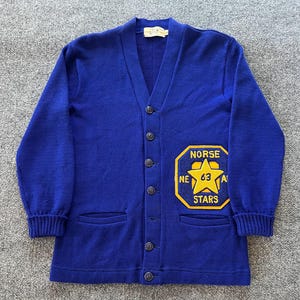 May include: A royal blue V-neck cardigan sweater featuring a large yellow patch on the lower right side. The patch is octagonal with a central star, the text "NORSE STARS", and the number "63". The sweater has long sleeves and dark buttons.