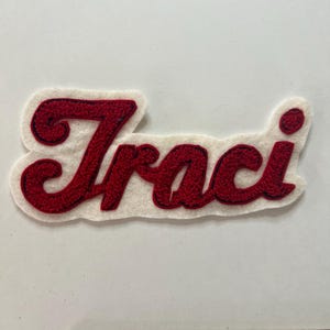 May include: A white felt patch featuring the name Traci embroidered in red cursive script with a dark outline. The patch is positioned above a wooden ruler, showing the name measures approximately 6 inches in total length from end to end.
