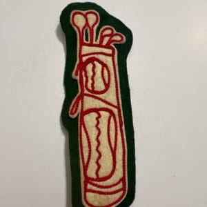 May include: A felt patch featuring a cream-colored golf bag with red embroidered outlines. The design includes golf clubs protruding from the top and a golf ball graphic on the front, all set against a dark green felt backing.