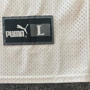 Op de afbeelding: Close-up van een witte mesh stof met een zwart rechthoekig label. Het label toont het witte PUMA-logo, de merknaam en de witte letter "L", die de maat aangeeft.