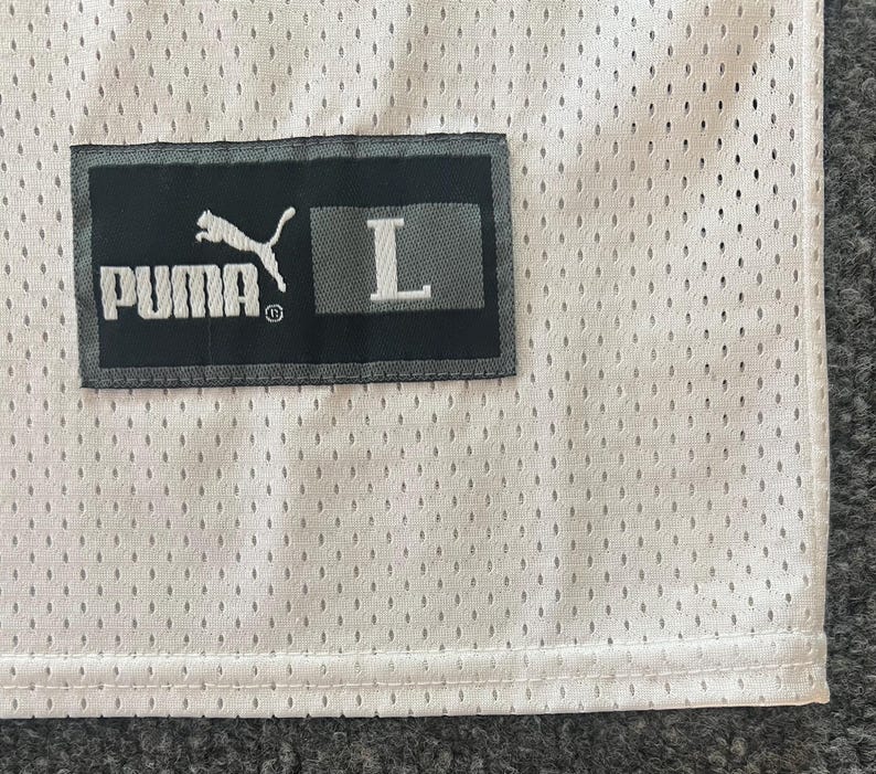Op de afbeelding: Close-up van een witte mesh stof met een zwart rechthoekig label. Het label toont het witte PUMA-logo, de merknaam en de witte letter "L", die de maat aangeeft.