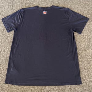 Op de afbeelding: Marineblauwe t-shirt met korte mouwen en het NFL-logo op de bovenrug. Het shirt is gemaakt van een zacht, ademend materiaal, perfect voor sportieve activiteiten of casual wear. Het heeft een klassieke ronde hals.