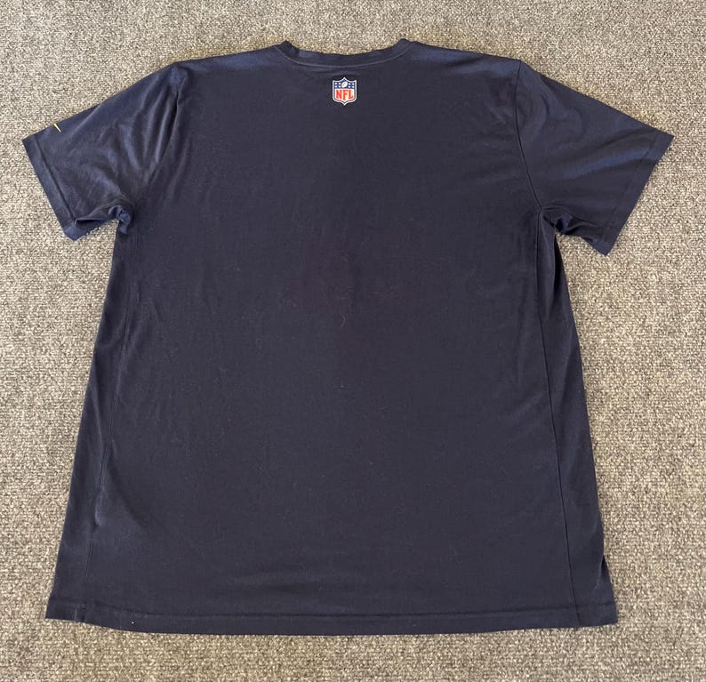 Op de afbeelding: Marineblauwe t-shirt met korte mouwen en het NFL-logo op de bovenrug. Het shirt is gemaakt van een zacht, ademend materiaal, perfect voor sportieve activiteiten of casual wear. Het heeft een klassieke ronde hals.