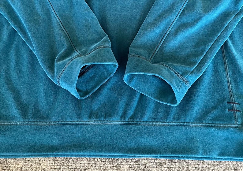 May include: A teal blue sweatshirt with a ribbed cuff and hem. The sweatshirt has a simple design and is made of a soft, comfortable fabric.