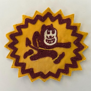 May include: A circular embroidered patch with a jagged, star-shaped yellow border. The center features a maroon-colored cartoon devil character with a white face, running toward the right, set against a solid yellow background.